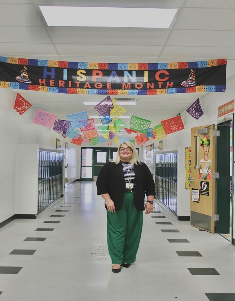 Mrs. Corcelles standing in the hallway with a banner saying Hispanic Heritage Month above her